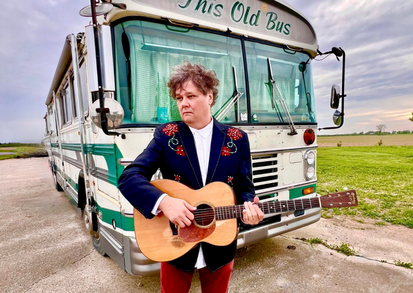 Artist Ron Sexsmith standing in front of old school bus with guitarx